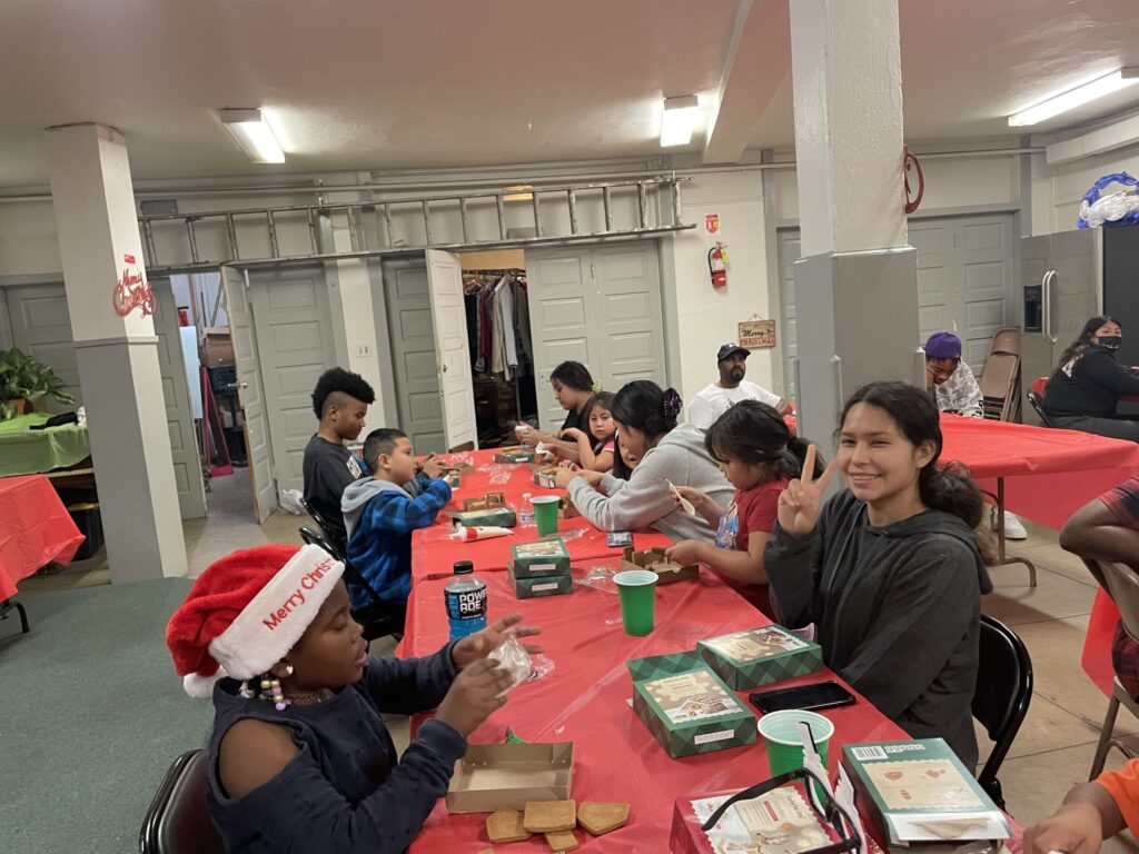Children and adults sit together at a long table during SBV’s 2023 Christmas party. The group enjoys festive activities with red tablecloths, gift boxes, and holiday hats in a brightly lit community room.